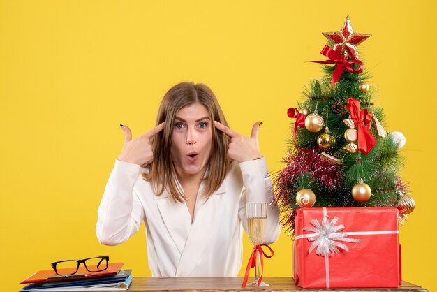 Vista frontal doctora sentada frente a su mesa sobre un fondo amarillo con árbol de navidad y cajas de regalo