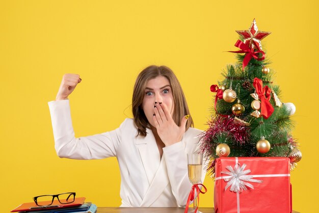 Vista frontal doctora sentada frente a su mesa sobre un fondo amarillo con árbol de navidad y cajas de regalo