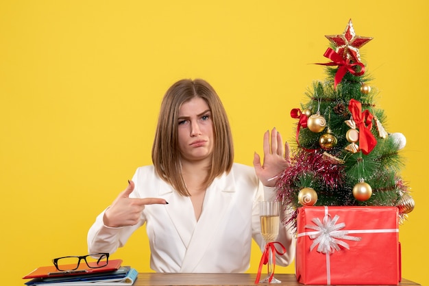 Vista frontal doctora sentada frente a su mesa sobre un fondo amarillo con árbol de navidad y cajas de regalo