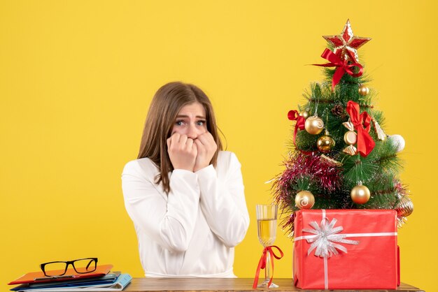 Vista frontal doctora sentada frente a su mesa sobre un fondo amarillo con árbol de navidad y cajas de regalo