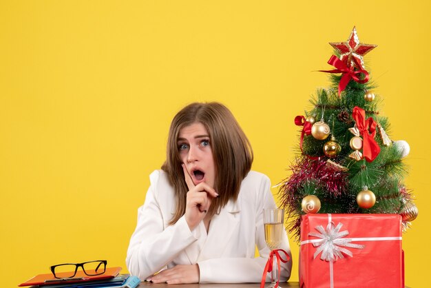 Vista frontal doctora sentada frente a su mesa sobre un fondo amarillo con árbol de navidad y cajas de regalo