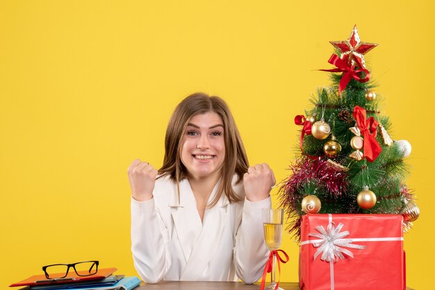 Vista frontal doctora sentada frente a su mesa sobre un fondo amarillo con árbol de navidad y cajas de regalo