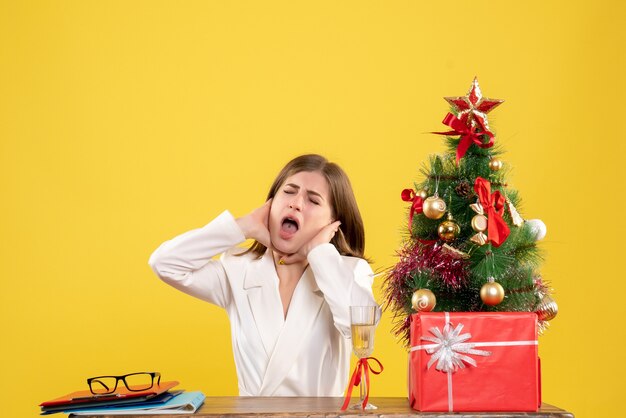 Vista frontal doctora sentada frente a su mesa sobre un fondo amarillo con árbol de navidad y cajas de regalo