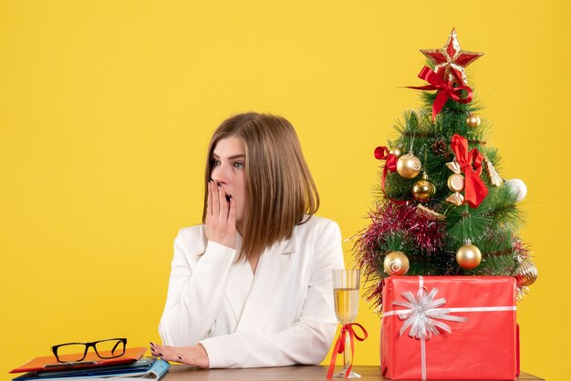 Vista frontal doctora sentada frente a su mesa sobre un fondo amarillo con árbol de navidad y cajas de regalo