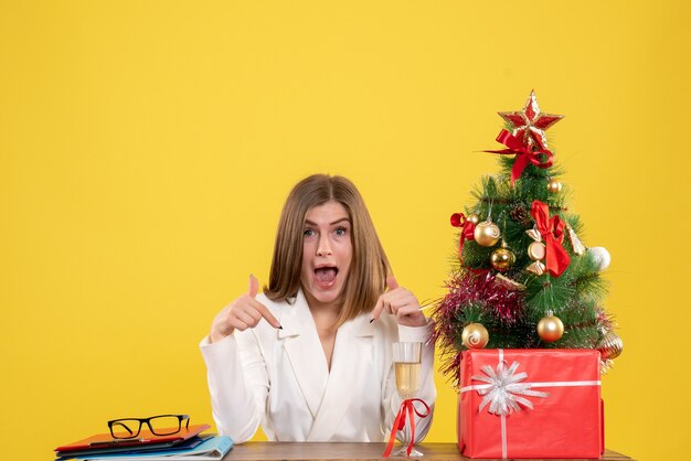 Vista frontal doctora sentada frente a su mesa sobre un fondo amarillo con árbol de navidad y cajas de regalo