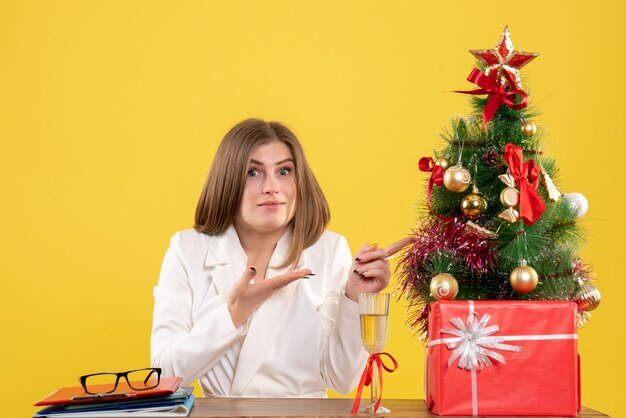 Vista frontal doctora sentada frente a su mesa sobre un fondo amarillo con árbol de navidad y cajas de regalo