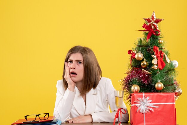 Vista frontal doctora sentada frente a su mesa sobre un fondo amarillo con árbol de navidad y cajas de regalo