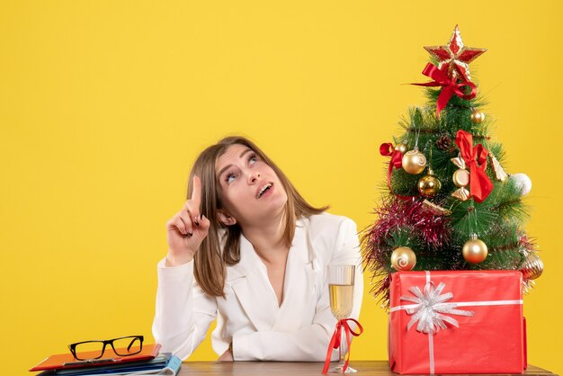 Vista frontal doctora sentada frente a su mesa sobre un fondo amarillo con árbol de navidad y cajas de regalo
