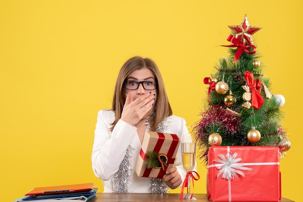 Vista frontal doctora sentada frente a su mesa sosteniendo presente en amarillo con árbol de navidad y cajas de regalo