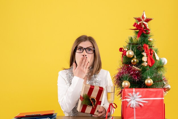 Vista frontal doctora sentada frente a su mesa sosteniendo presente sobre fondo amarillo con árbol de navidad y cajas de regalo