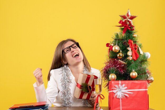 Vista frontal doctora sentada frente a su mesa sosteniendo presente sobre fondo amarillo con árbol de navidad y cajas de regalo