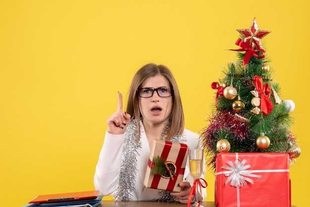 Vista frontal doctora sentada frente a su mesa sosteniendo presente sobre fondo amarillo con árbol de navidad y cajas de regalo