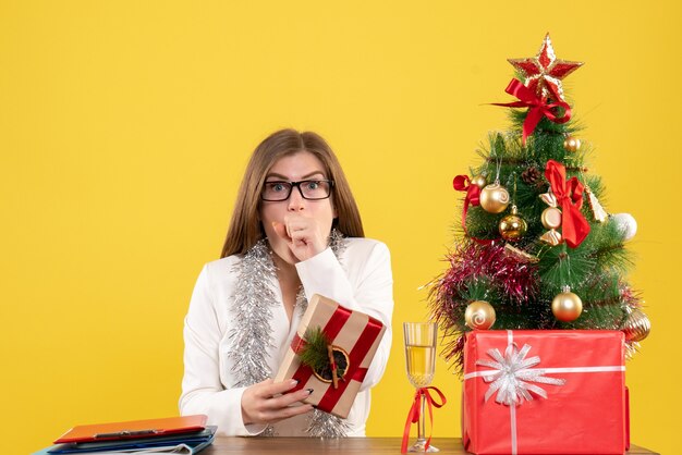 Vista frontal doctora sentada frente a su mesa sosteniendo presente sobre fondo amarillo con árbol de navidad y cajas de regalo