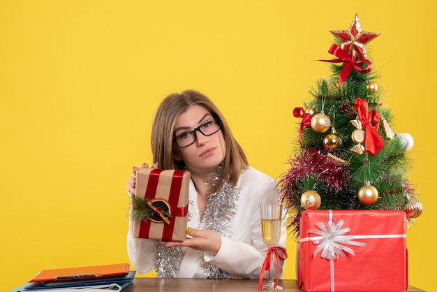 Vista frontal doctora sentada frente a su mesa sosteniendo presente sobre fondo amarillo con árbol de navidad y cajas de regalo