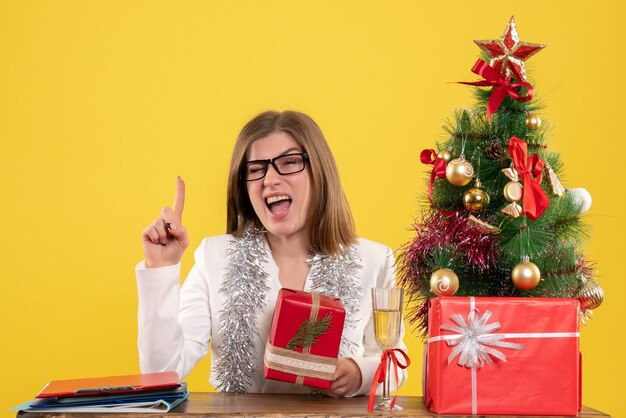 Vista frontal doctora sentada frente a su mesa sosteniendo presente sobre fondo amarillo con árbol de navidad y cajas de regalo