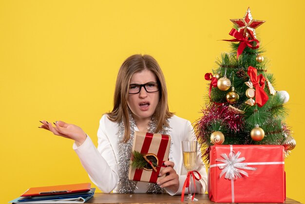 Vista frontal doctora sentada frente a su mesa sosteniendo presente sobre fondo amarillo con árbol de navidad y cajas de regalo