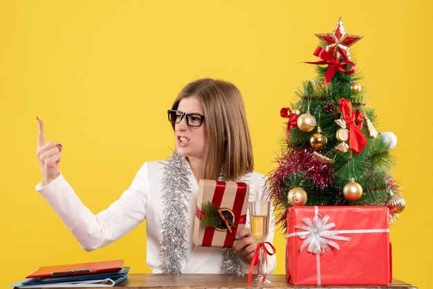 Vista frontal doctora sentada frente a su mesa sosteniendo presente sobre fondo amarillo con árbol de navidad y cajas de regalo