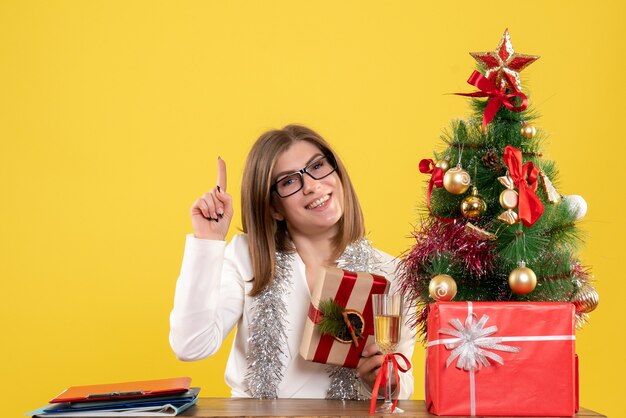 Vista frontal doctora sentada frente a su mesa sosteniendo presente sobre fondo amarillo con árbol de navidad y cajas de regalo