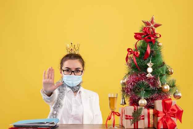 Vista frontal doctora sentada en máscara estéril pidiendo detenerse sobre fondo amarillo con árbol de navidad y cajas de regalo