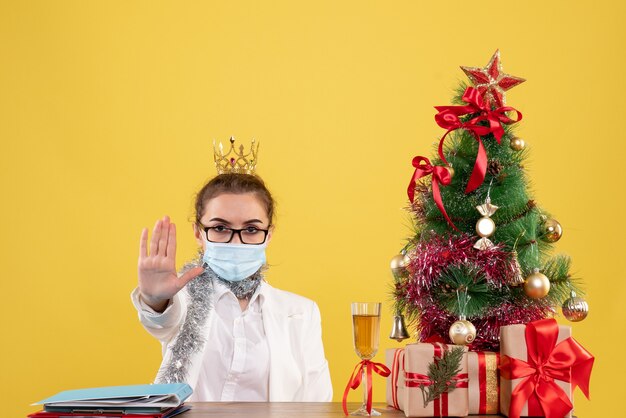 Vista frontal doctora sentada en máscara estéril pidiendo detenerse sobre fondo amarillo con árbol de navidad y cajas de regalo