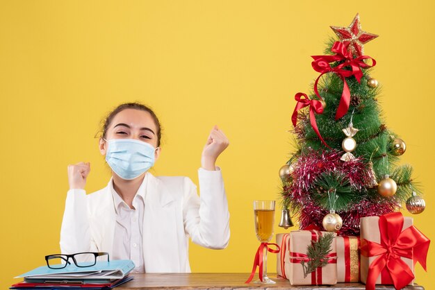 Vista frontal doctora sentada en máscara estéril sobre fondo amarillo con árbol de navidad y cajas de regalo