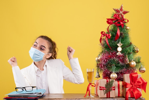 Vista frontal doctora sentada en máscara estéril sobre fondo amarillo con árbol de navidad y cajas de regalo