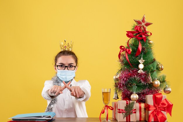 Vista frontal doctora sentada en máscara estéril sobre fondo amarillo con árbol de navidad y cajas de regalo