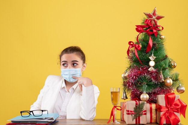 Vista frontal doctora sentada en máscara estéril sobre fondo amarillo con árbol de navidad y cajas de regalo