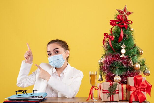 Vista frontal doctora sentada en máscara estéril sobre fondo amarillo con árbol de navidad y cajas de regalo