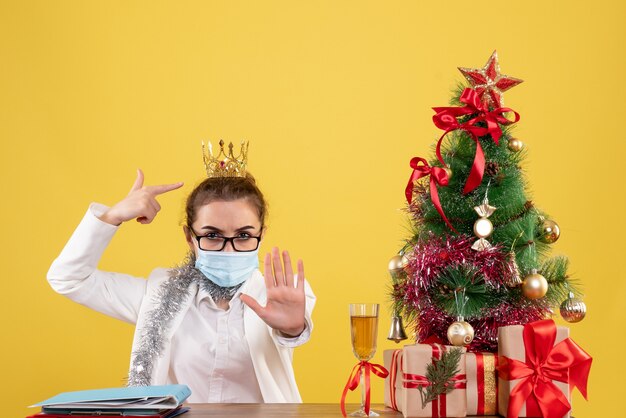 Vista frontal doctora sentada en máscara estéril sobre fondo amarillo con árbol de navidad y cajas de regalo