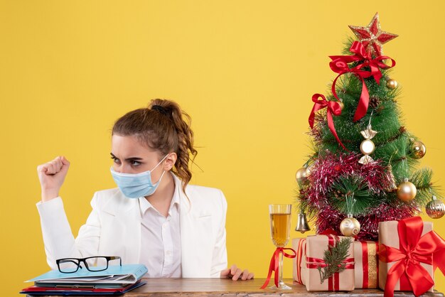 Vista frontal doctora sentada en máscara estéril sobre fondo amarillo con árbol de navidad y cajas de regalo