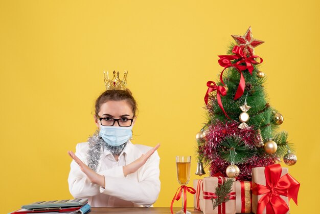 Vista frontal doctora sentada en máscara estéril sobre fondo amarillo con árbol de navidad y cajas de regalo