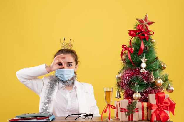 Vista frontal doctora sentada en máscara estéril sobre fondo amarillo con árbol de navidad y cajas de regalo