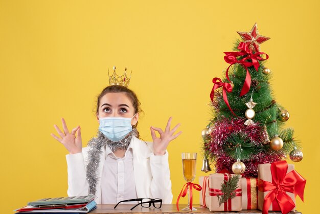 Vista frontal doctora sentada en máscara estéril sobre fondo amarillo con árbol de navidad y cajas de regalo