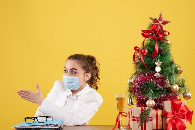 Vista frontal doctora sentada en máscara protectora sobre fondo amarillo con árbol de navidad y cajas de regalo