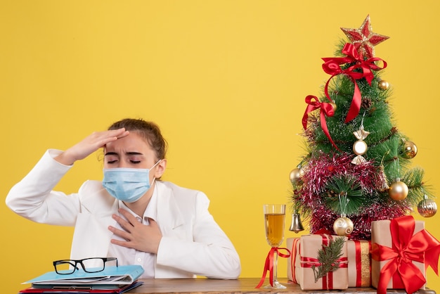 Vista frontal doctora sentada en máscara protectora sobre fondo amarillo con árbol de navidad y cajas de regalo