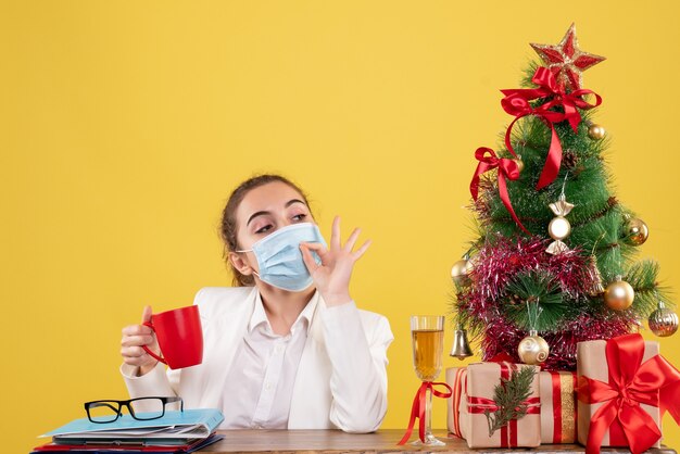 Vista frontal doctora sentada en máscara protectora sobre fondo amarillo con árbol de navidad y cajas de regalo