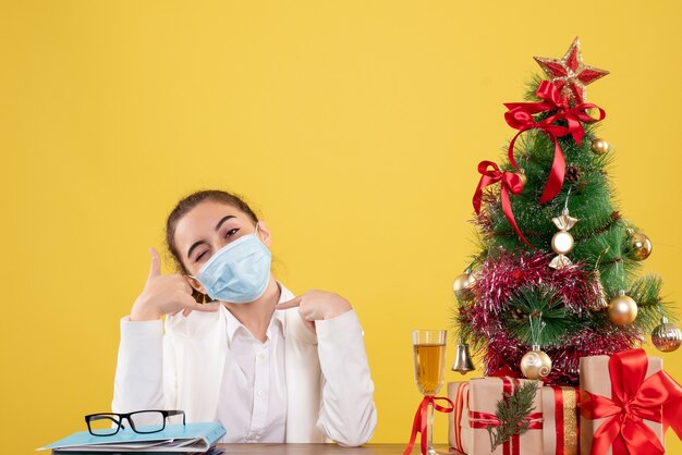 Vista frontal doctora sentada en máscara protectora sobre fondo amarillo con árbol de navidad y cajas de regalo