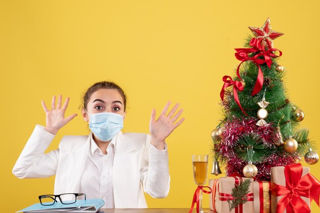 Vista frontal doctora sentada en máscara protectora sobre fondo amarillo con árbol de navidad y cajas de regalo