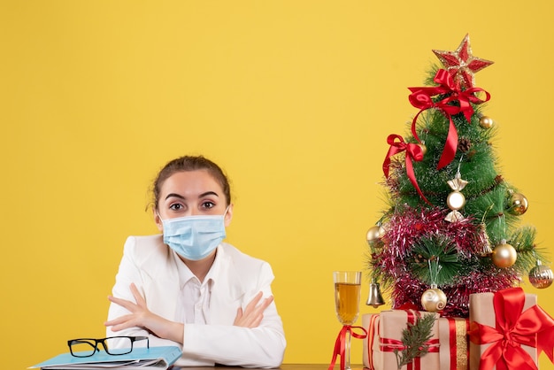 Vista frontal doctora sentada en máscara protectora sobre fondo amarillo con árbol de navidad y cajas de regalo