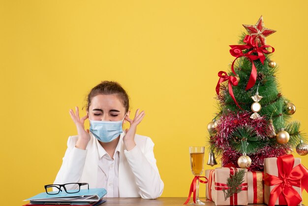 Vista frontal doctora sentada en máscara protectora sobre fondo amarillo con árbol de navidad y cajas de regalo