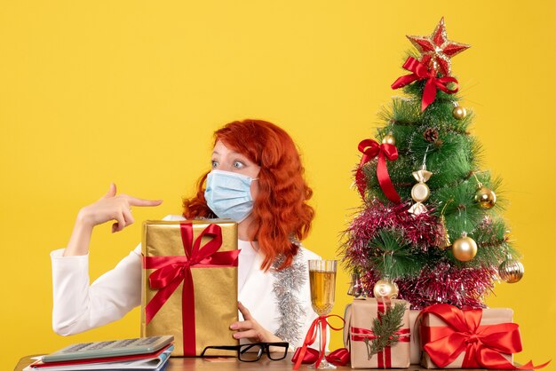 Vista frontal doctora sentada en máscara con regalos de Navidad y árbol sobre fondo amarillo