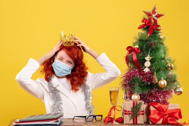 Vista frontal doctora sentada en máscara con regalos de Navidad con corona sobre fondo amarillo