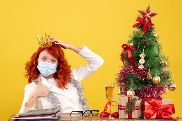 Vista frontal doctora sentada en máscara con regalos de Navidad con corona sobre fondo amarillo