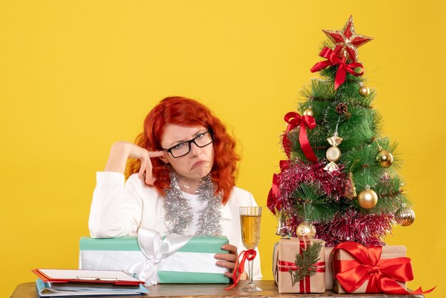 Vista frontal doctora sentada con regalos de navidad y árbol en escritorio amarillo