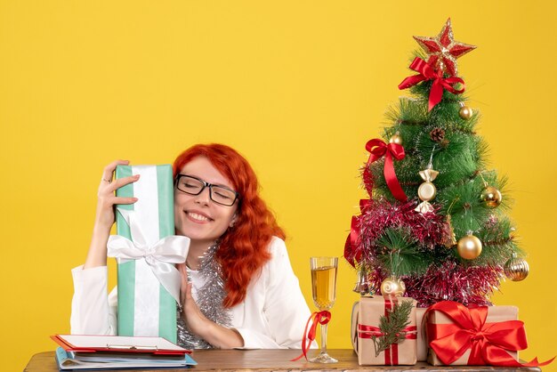 Vista frontal doctora sentada con regalos de Navidad y árbol sobre fondo amarillo