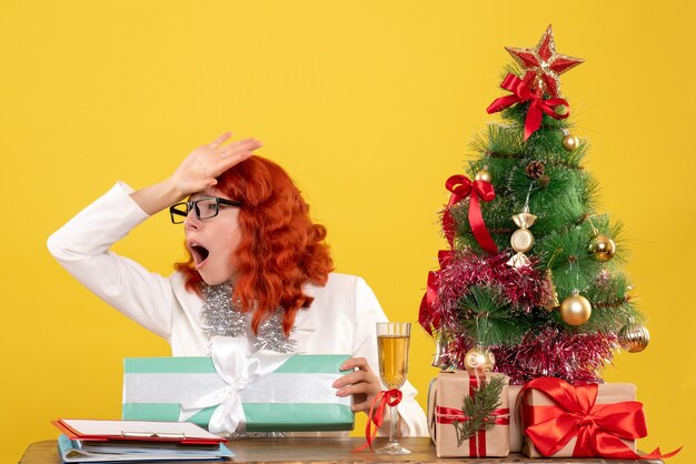 Vista frontal doctora sentada con regalos de Navidad y árbol sobre fondo amarillo