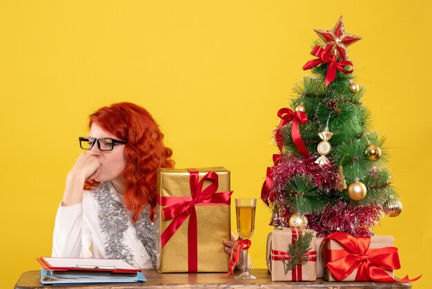 Vista frontal doctora sentada con regalos de Navidad y árbol sobre fondo amarillo