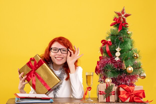 Vista frontal doctora sentada con regalos de Navidad y árbol sobre fondo amarillo
