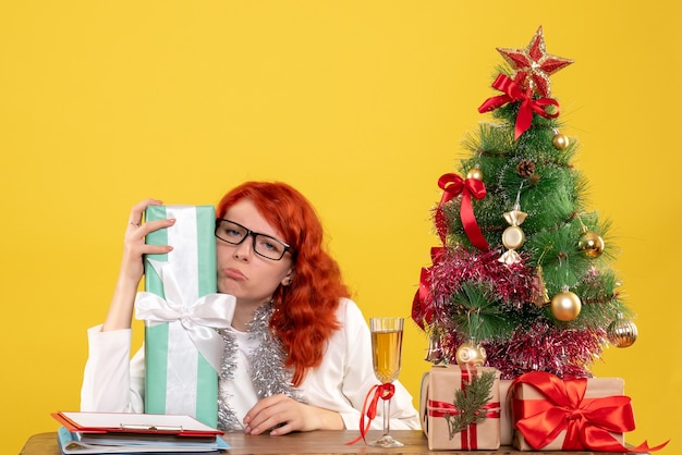 Vista frontal doctora sentada con regalos de Navidad y árbol sobre fondo amarillo